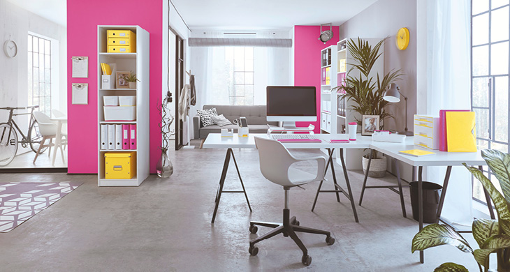 Blick in ein loftartiges Wohnzimmer: Im Vordergrund steht ein weißer Schreibtisch mit weißem Drehstuhl und Büromaterial von Leitz, im Hintergrund sieht man ein Wohnzimmer mit pinken Wänden und Regalen, die mit Ordnern und Boxen von Leitz bestückt sind.