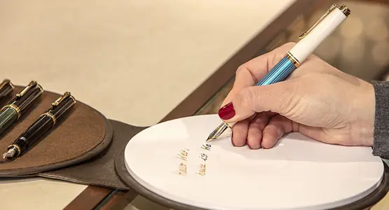 Eine Hand schreibt mit Füllhalter auf ein rundes Stück Papier. Daneben sind weitere Schreibgeräte im Anschnitt zu sehen.