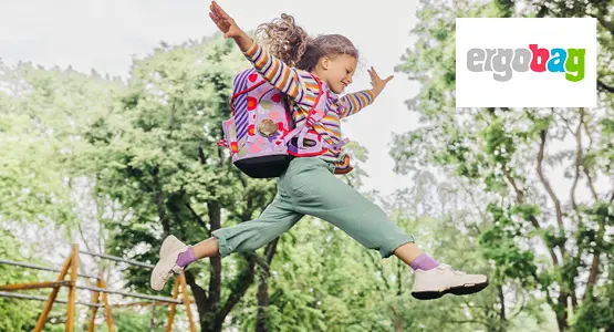 Vor Baumen springt ein Kind mit Schulranzen auf dem Rücken in die Luft.