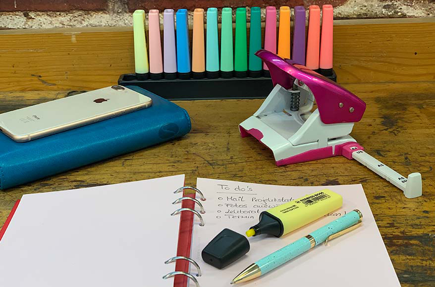 Man sieht einen bunt eingerichteten Arbeitsplatz. Im Hintergrund stehen zahlreiche Textmarker der Marke Stabilo, ein rosafarbenen Locher sowie ein Planer von Filofax auf dem ein Smartphone liegt. Im Vordergrund liegt ein Notizbuch der Marke Filofax, in dem eine to-do-Liste geführt wird, sowie ein türkiser Kugelschreiber